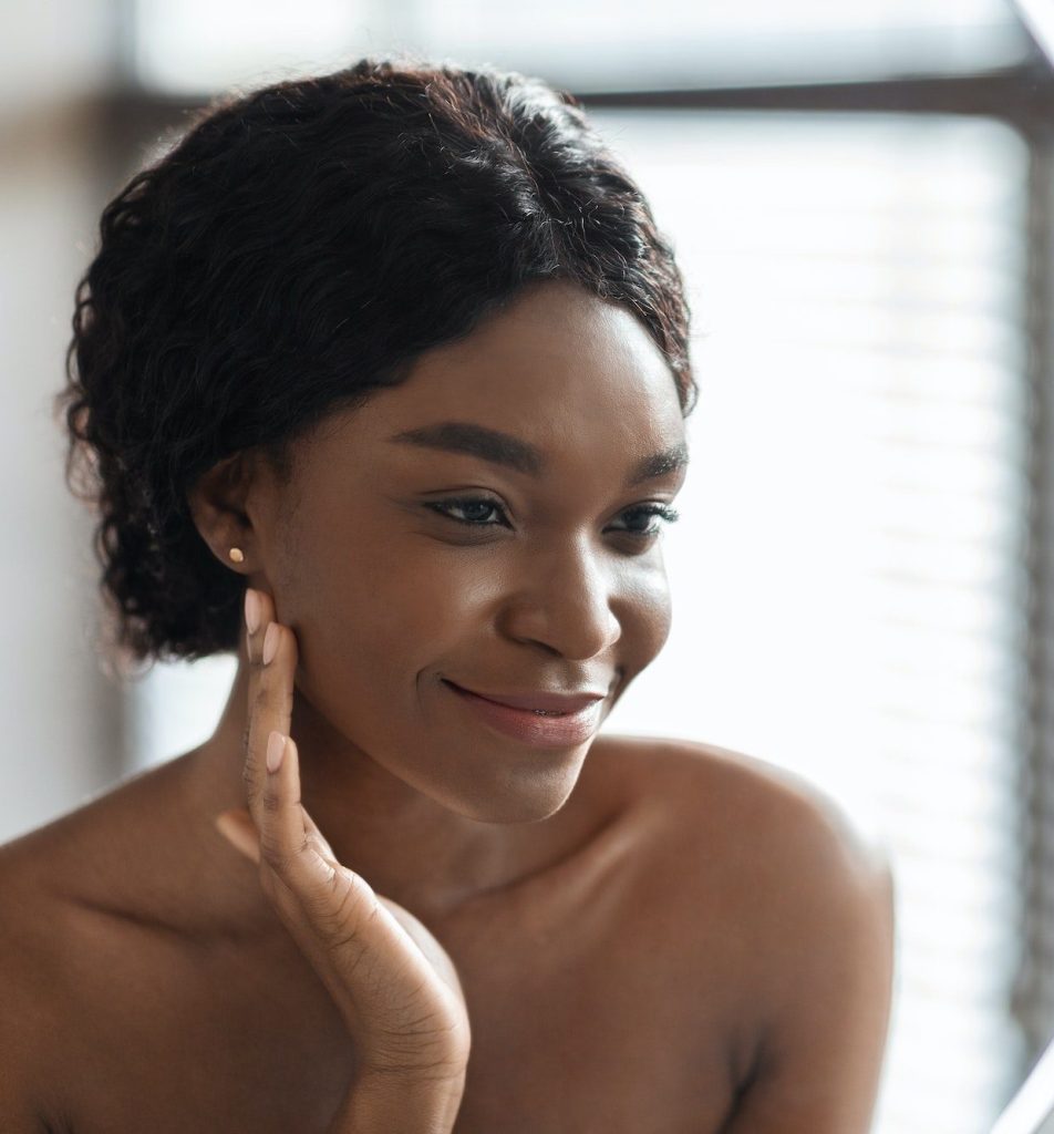 Mirror reflection of pretty black woman applying facial product Facial Harmonization