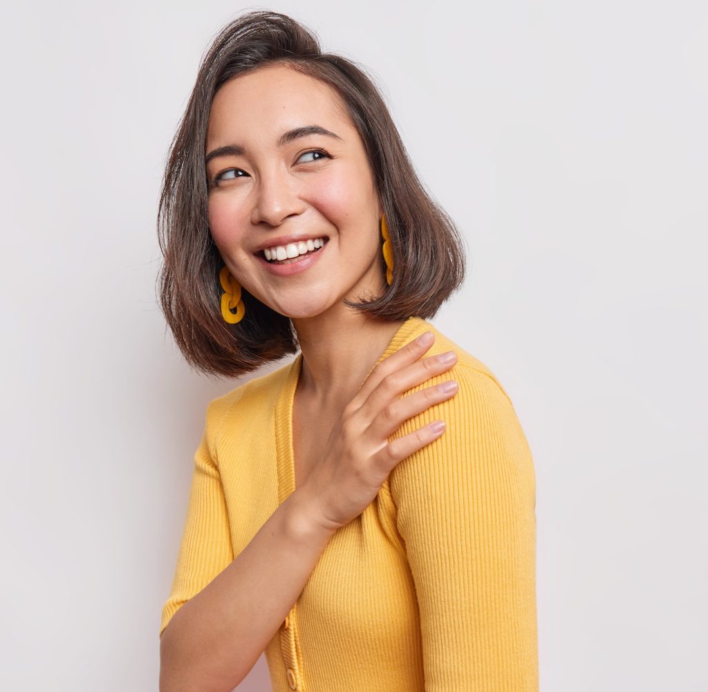 Sideways shot of sincere happy woman with eastern appearance wears casual yellow jumper poses agains