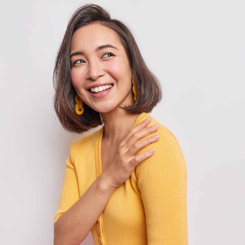 Sideways shot of sincere happy woman with eastern appearance wears casual yellow jumper poses agains